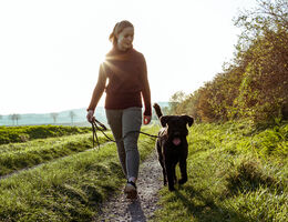 A woman walks a dog along a dirt trail.